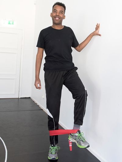 En man står vid en vägg med ett gummiband runt  fotlederna. Han håller sig i väggen och lyfter ett ben.