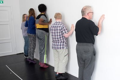 Fem personer står på tå med ansiktena mot en vägg och håller fingertoppar mot väggen.